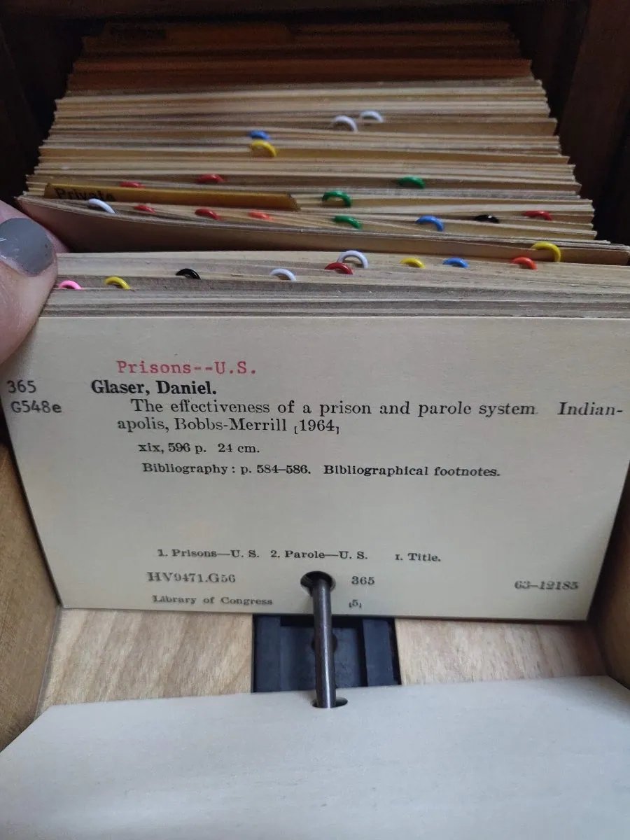 An open library card catalogue drawer packed with index cards. One card is raised to show a bibliographic reference, with coloured clips marking groups.