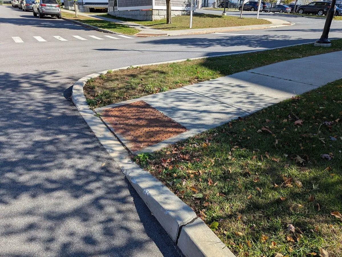 A long gentle pavement ramp leading down to a lowered kerb at a road, with a strip of bumpy tactile paving before the edge.