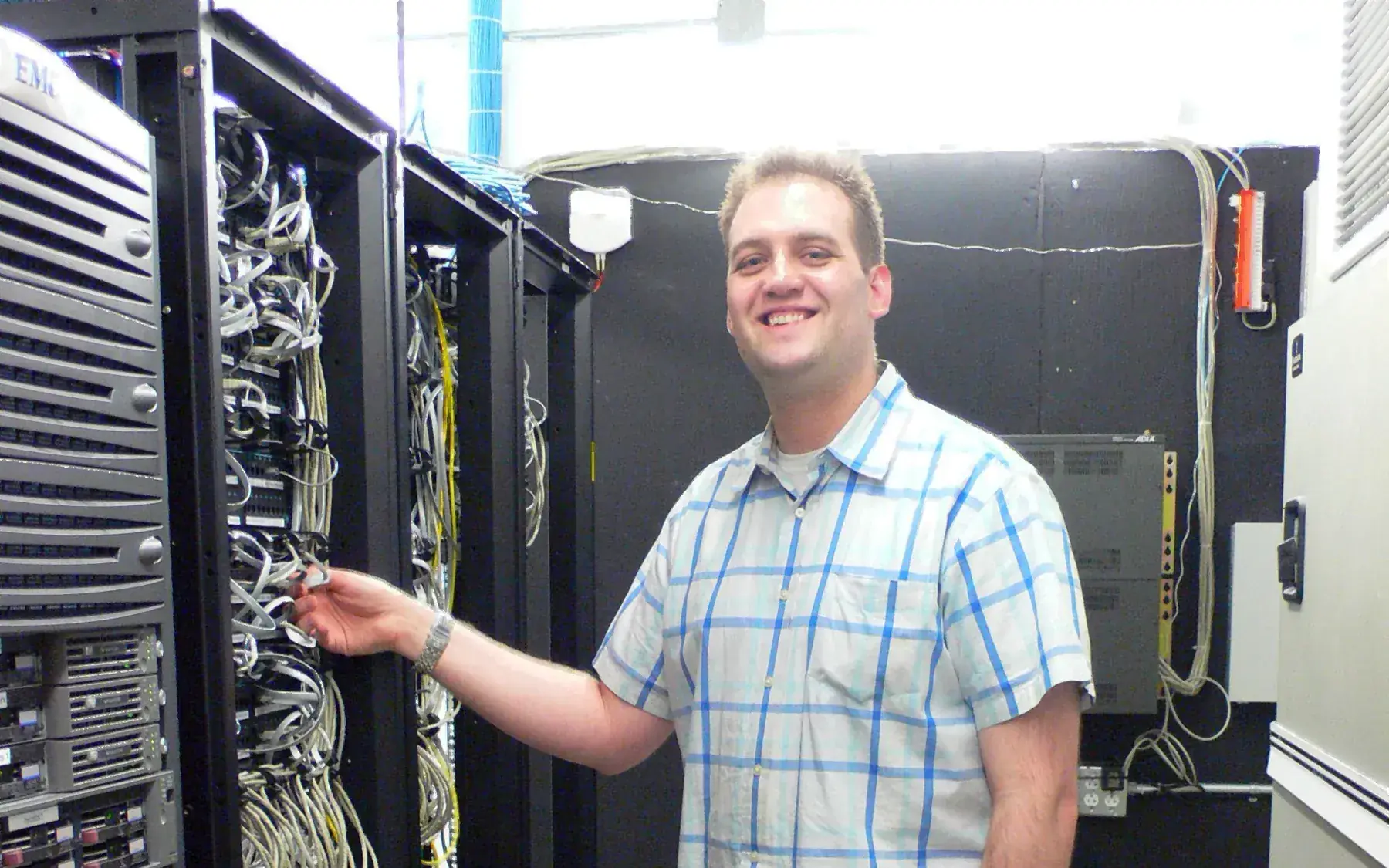 A system administrator standing at an open server rack, working among cabled network equipment in a data center aisle.