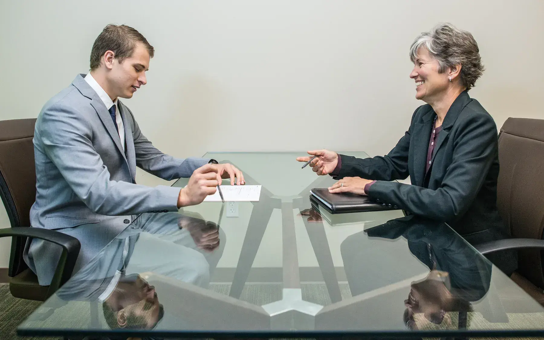 A young man sits across a desk from a mature woman during a job interview, mid-conversation.
