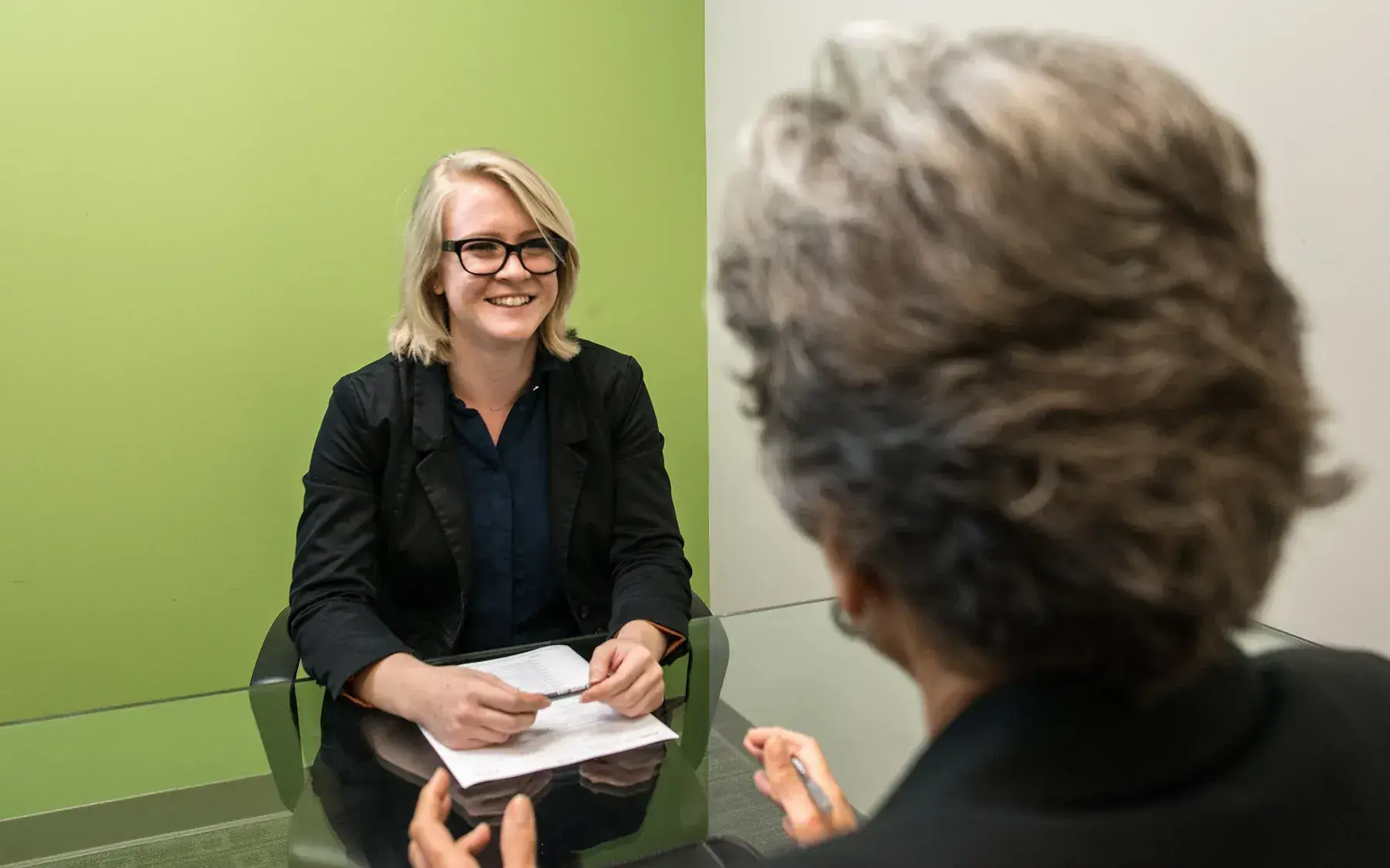 A smiling young woman talks with a mature interviewer seated opposite her at an office desk.